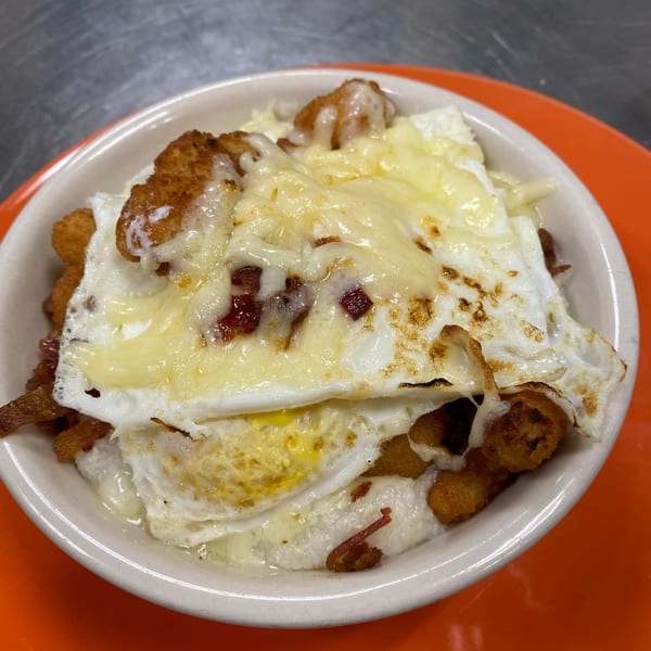 A diner breakfast bowl with grits, fried eggs, melted cheese, bacon bits, and tater tots.