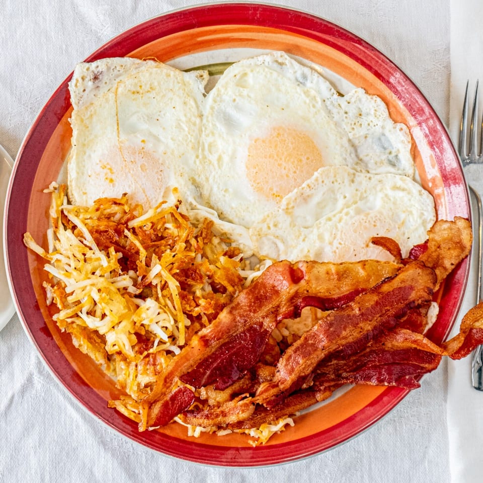A diner breakfast plate with three fried eggs, crispy hash browns, and several strips of bacon.